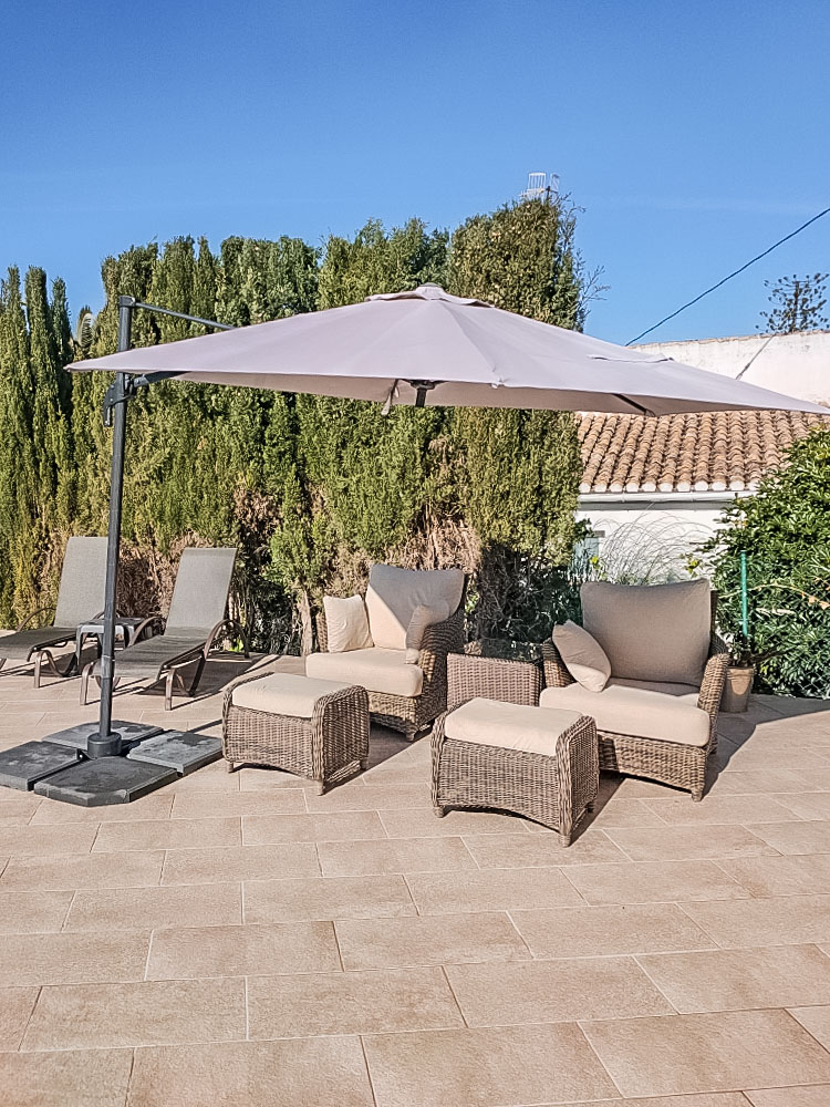 Buitenlounge met grijze rieten fauteuils, voetenbankjes en een grote zweefparasol op een betegeld terras, omgeven door volgroeide hagen.