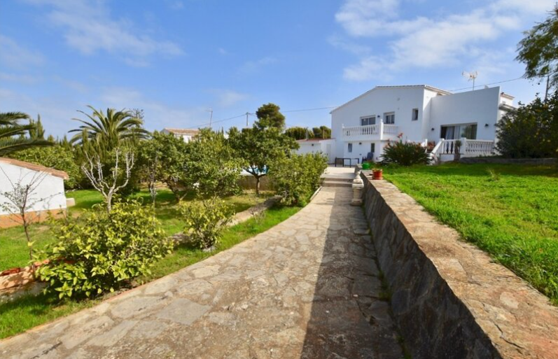 Een natuurlijk stenen pad leidt door de mediterrane tuin naar de witte villa, omgeven door gazons, palmbomen en klassieke witte balustrades.