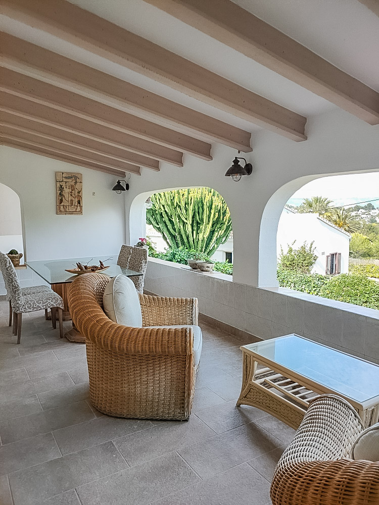Ruime overdekte naya met houten balkenplafond, rieten meubilair en traditionele bogen met uitzicht op de mediterrane tuin.
