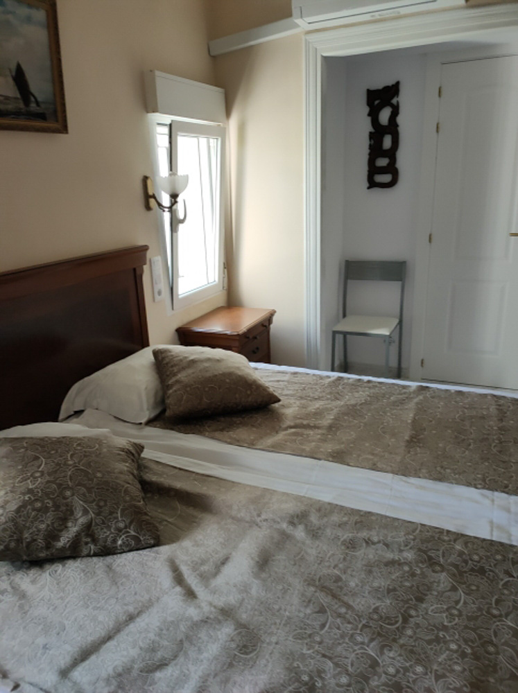 Slaapkamer met tweepersoonsbed, klassiek houten hoofdbord en airconditioning. Voorzien van natuurlijk licht via een zijraam, een nachtkastje en een praktische stoel in de hoek.