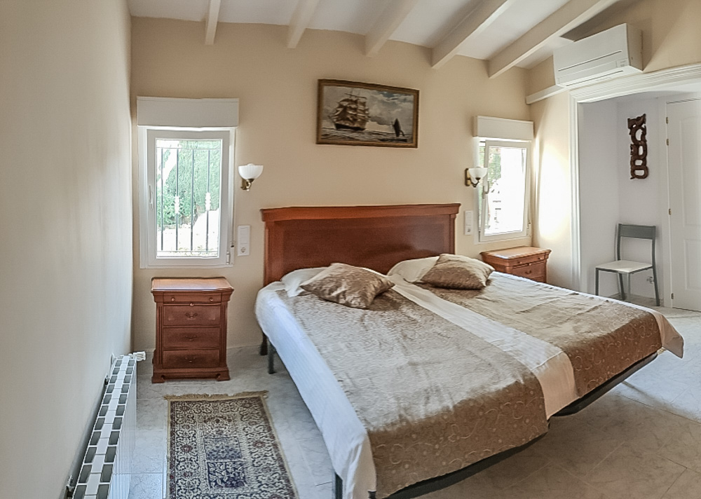 Slaapkamer met wit balkenplafond, houten tweepersoonsbed met bijpassende nachtkastjes, airconditioning en radiatorverwarming.