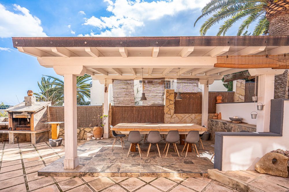 Buiten-eethoek onder een witte houten pergola met een robuuste houten tafel, moderne stoelen en een stenen barbecue op het betegelde terras.