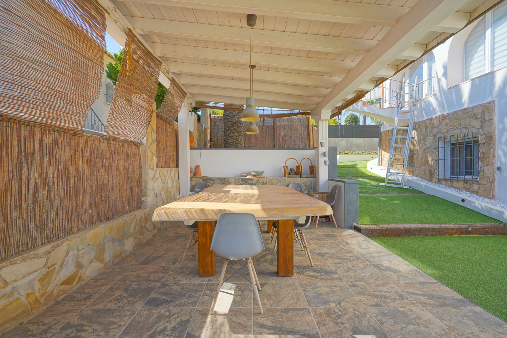 Buiten eethoek onder een witte houten pergola, voorzien van een massief houten tafel, tegelvloer en privacyschermen grenzend aan het gazon.