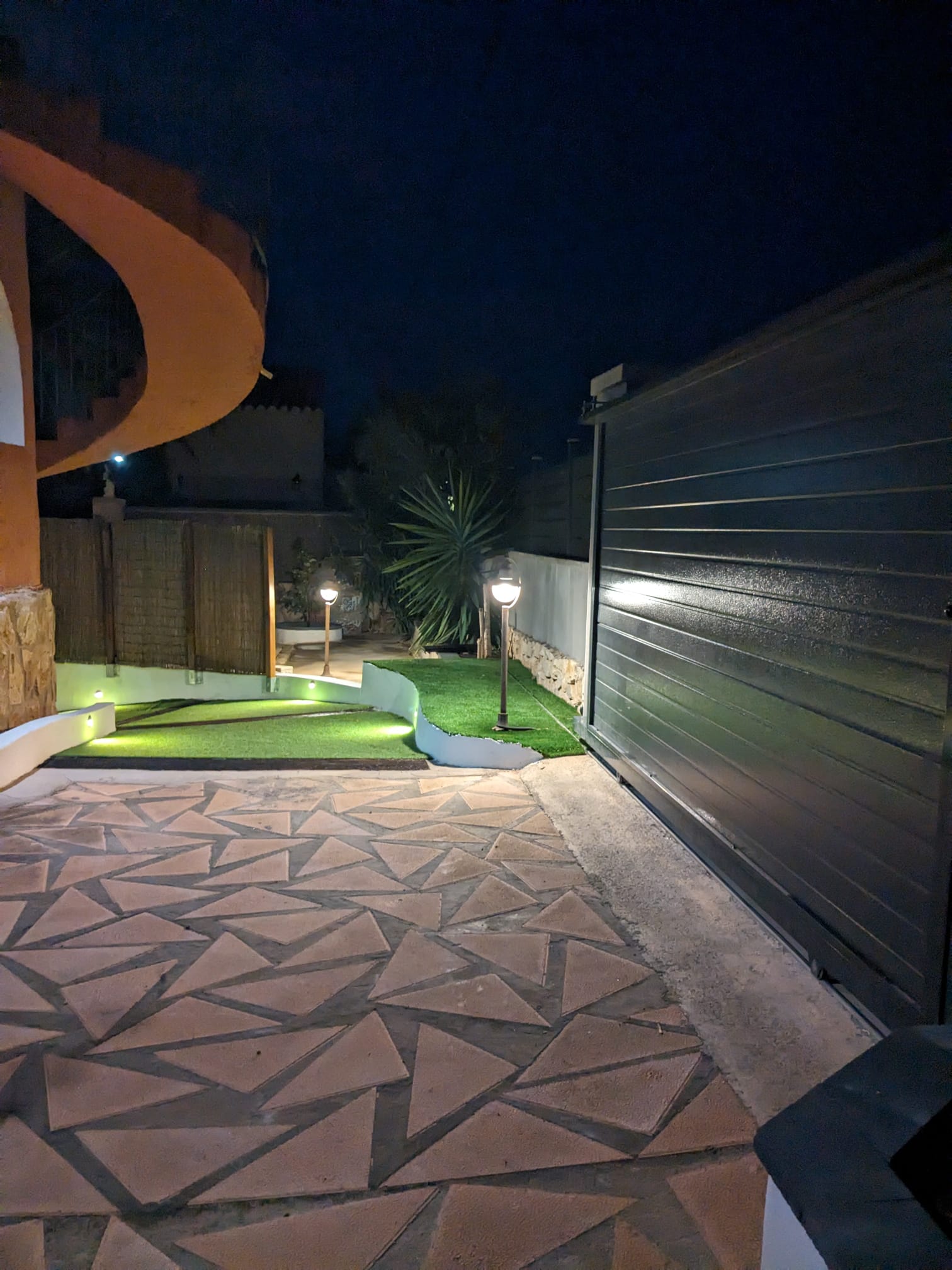 Buitenentree bij nacht met grijze schuifpoort, geometrische bestrating en geïntegreerde verlichting langs het kunstgras en de muren.