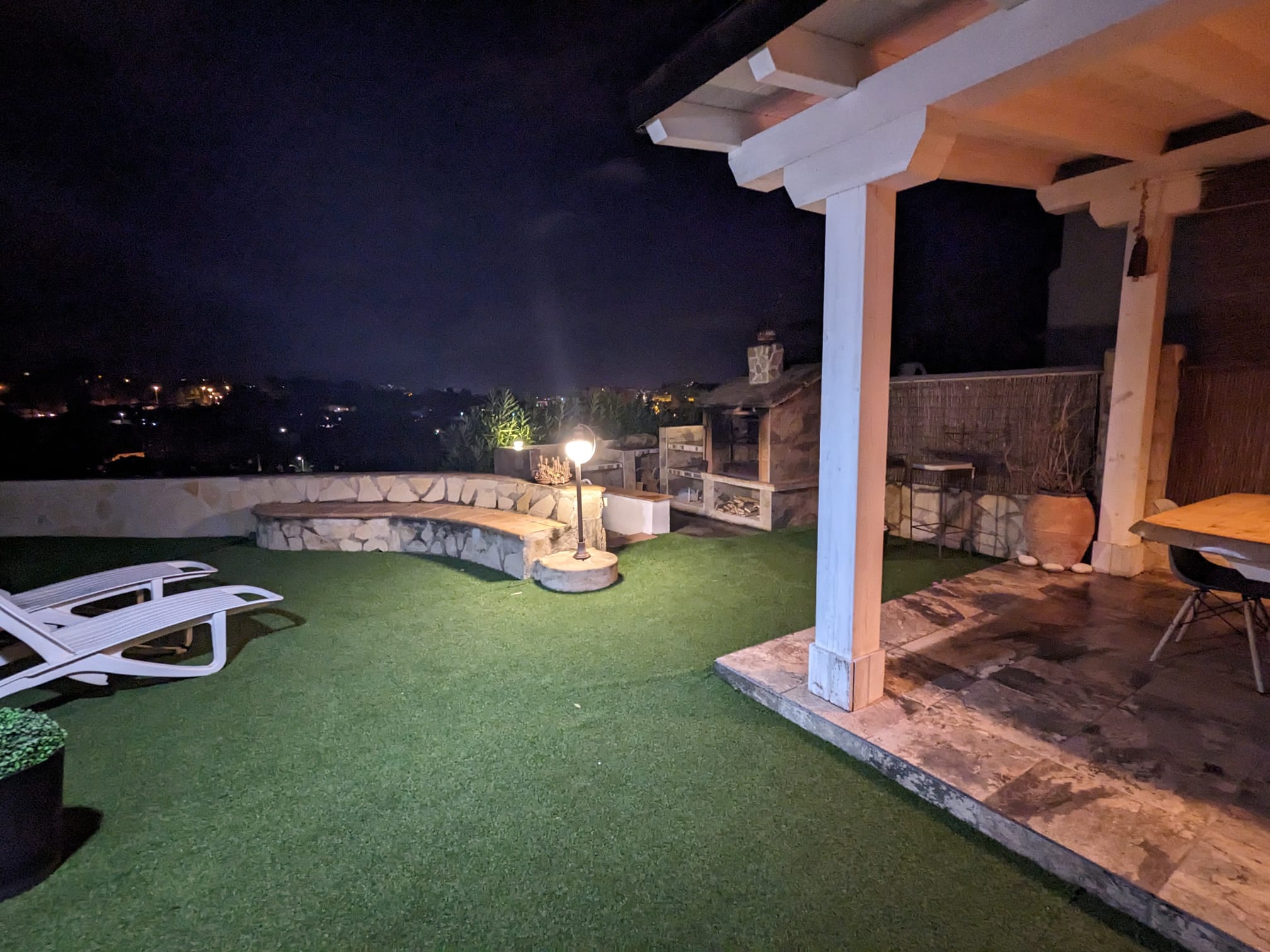 Buitenruimte bij nacht met een stenen barbecue, gebogen gemetselde bank en houten pergola met eethoek naast een kunstgrasveld met ligstoelen.