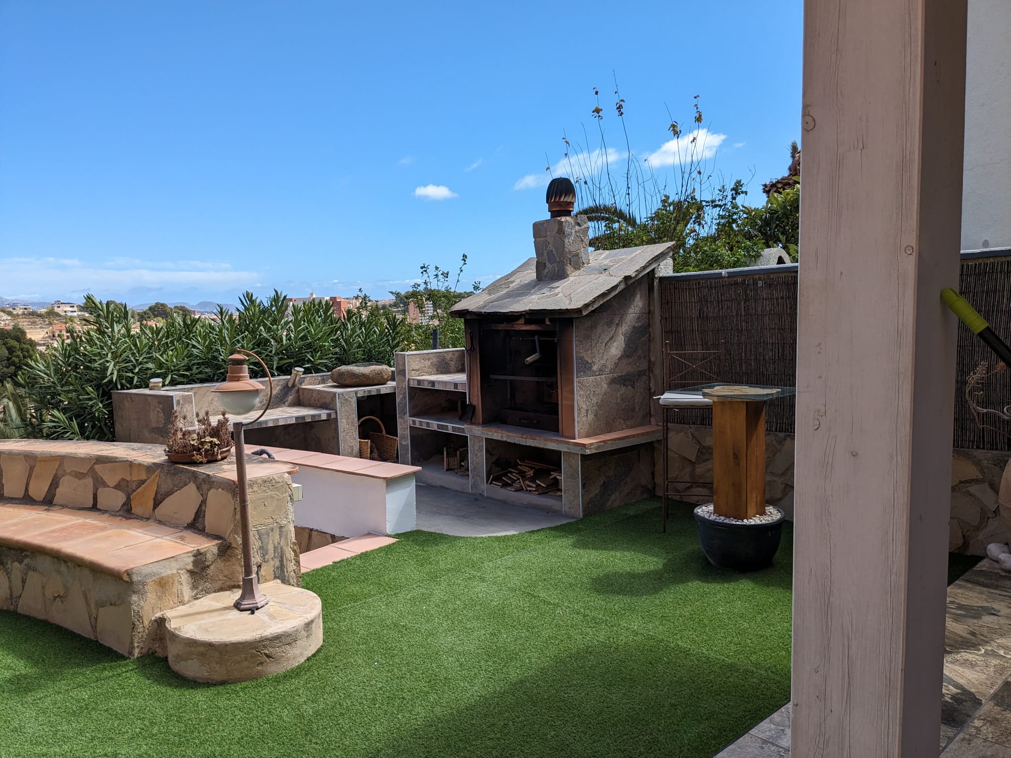 Buitenruimte met een natuurstenen barbecue, ingebouwde werkbladen, een gemetselde bank met terracotta tegels en een strak kunstgrasveld.