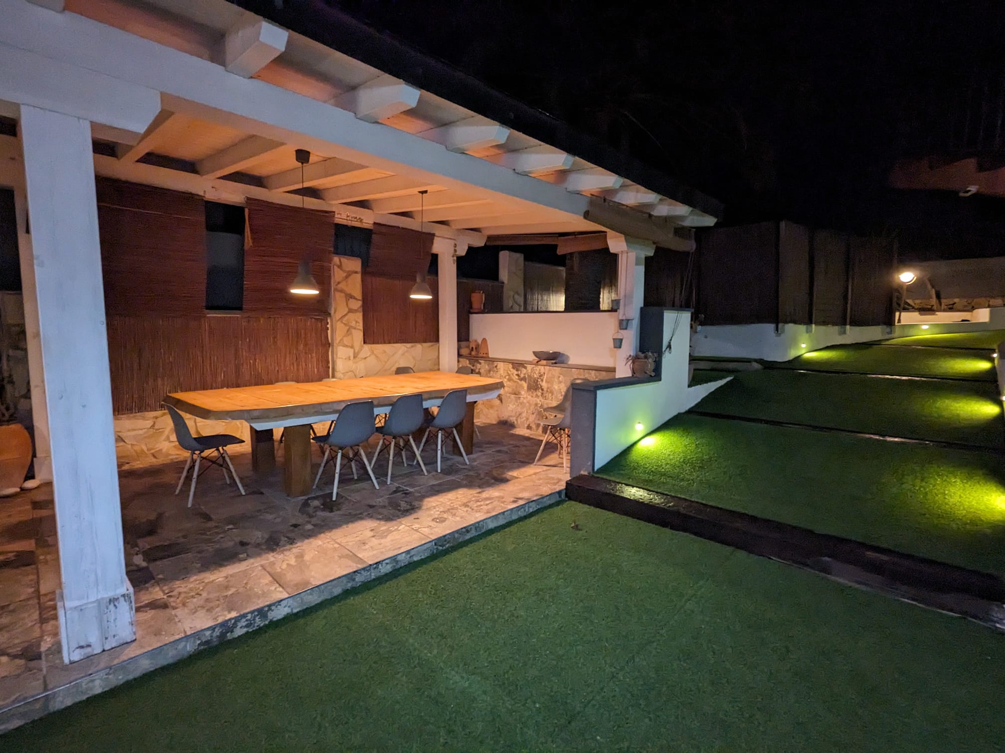 Buitenruimte met eethoek onder een witte houten pergola, voorzien van een robuuste houten tafel en stenen vloer naast een verlicht gazon met kunstgras.