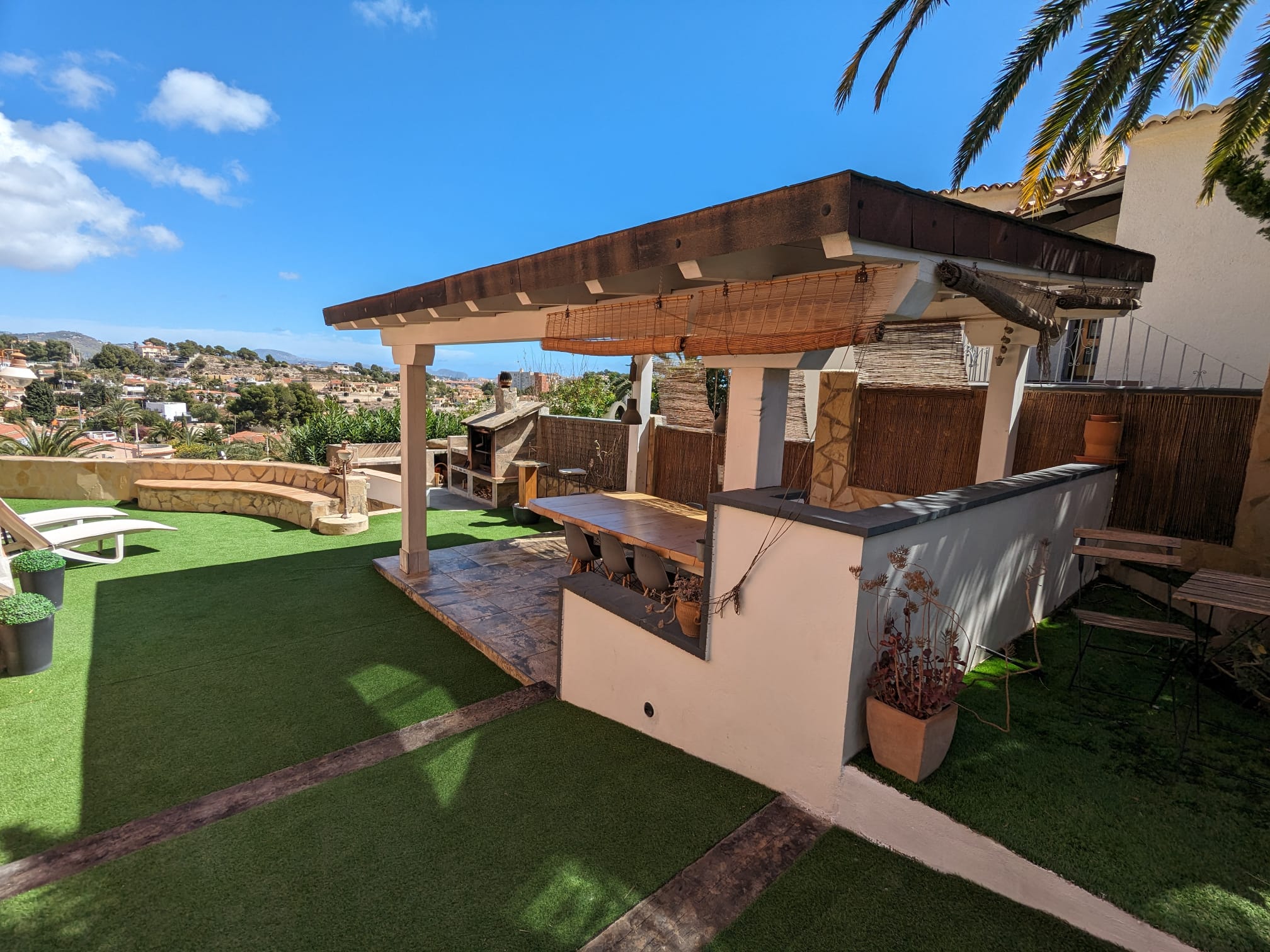 Buitenruimte met houten pergola, eethoek en stenen barbecue, omgeven door kunstgras en met weids uitzicht over de omgeving van Calpe.