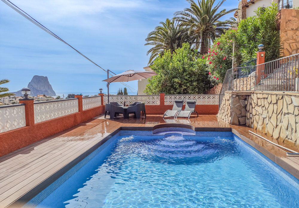 Buitenterras met blauw mozaïekzwembad, houten vlonder en uitzicht op de Peñón de Ifach. Voorzien van een eethoek met parasol en ligbedden.
