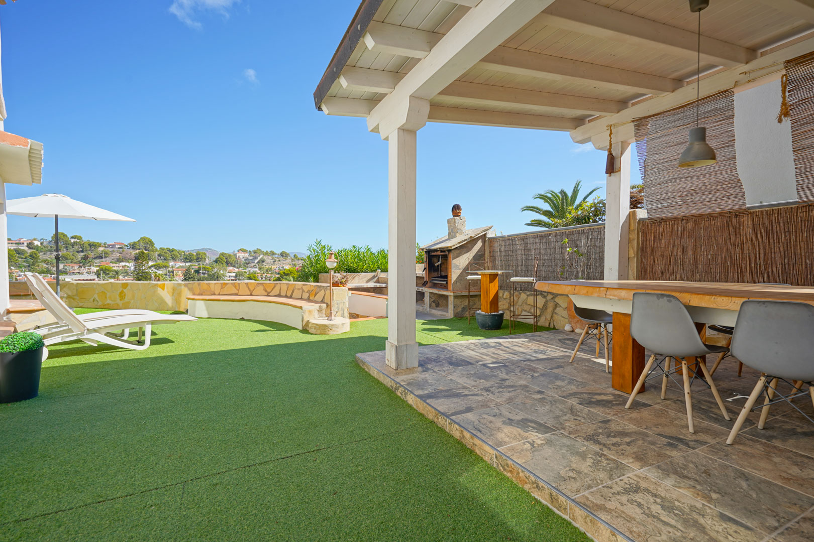Buitenterras met witte houten pergola, eethoek, stenen barbecue en kunstgrasveld met ligstoelen en vrij uitzicht over de omgeving.