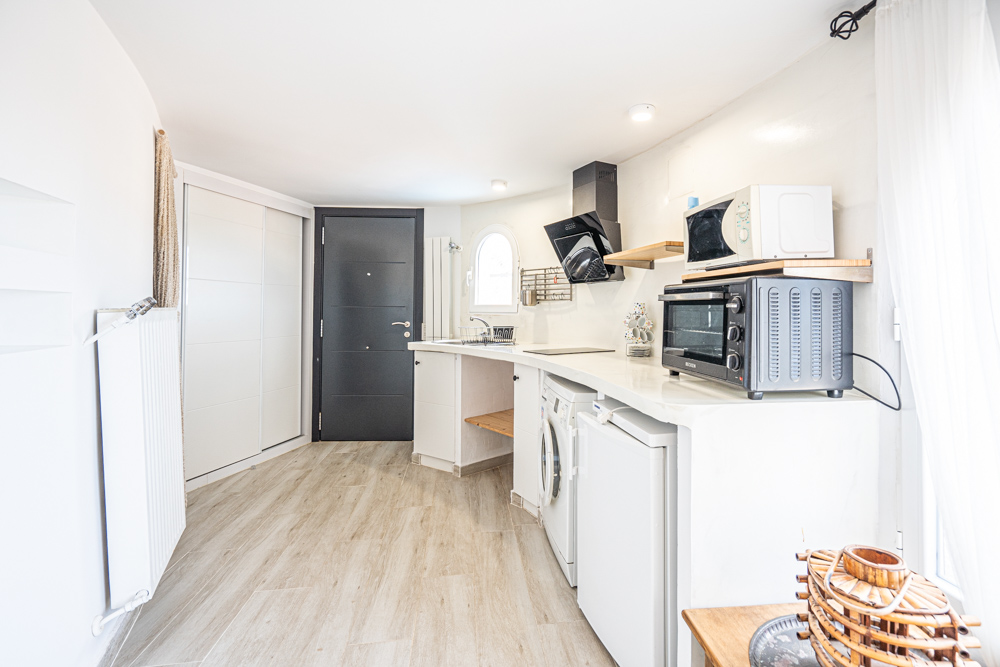 Compacte gerenoveerde keuken met een gebogen wit werkblad, moderne apparatuur, houtlook vloertegels en een eigen toegangsdeur.