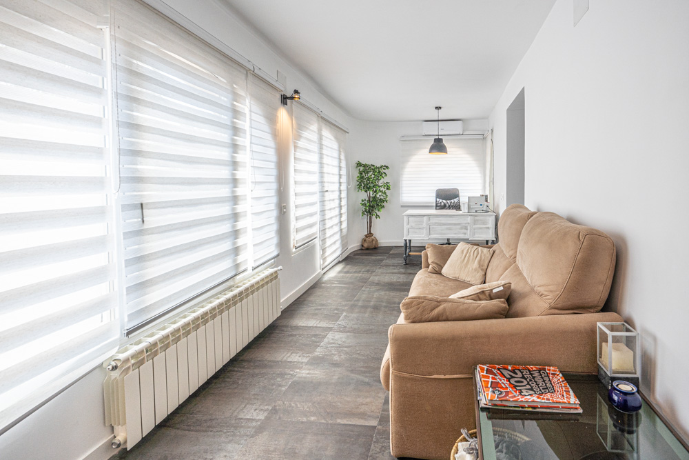 Lichte naya met donkere tegelvloer, beige bank en een werkplek. Voorzien van een radiator, airconditioning en grote ramen met witte duo-rolgordijnen.