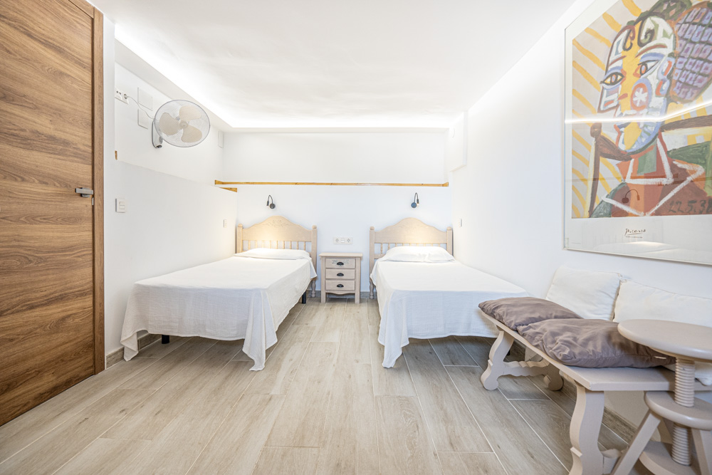 Lichte slaapkamer met twee eenpersoonsbedden, keramische vloer met houtlook, indirecte LED-plafondverlichting en een wandventilator.