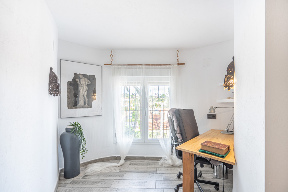 Lichte werkruimte met houten bureau, keramische tegels met houtlook en een raam met beveiligingsroosters voor natuurlijk daglicht.