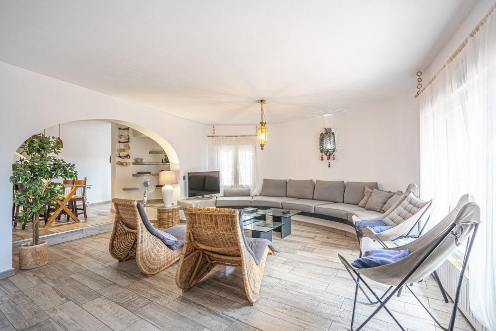 Lichte woonkamer met een ingebouwde ronde bank, rotan fauteuils en houtlook tegels. Een karakteristieke boog biedt doorgang naar de aangrenzende eetkamer.