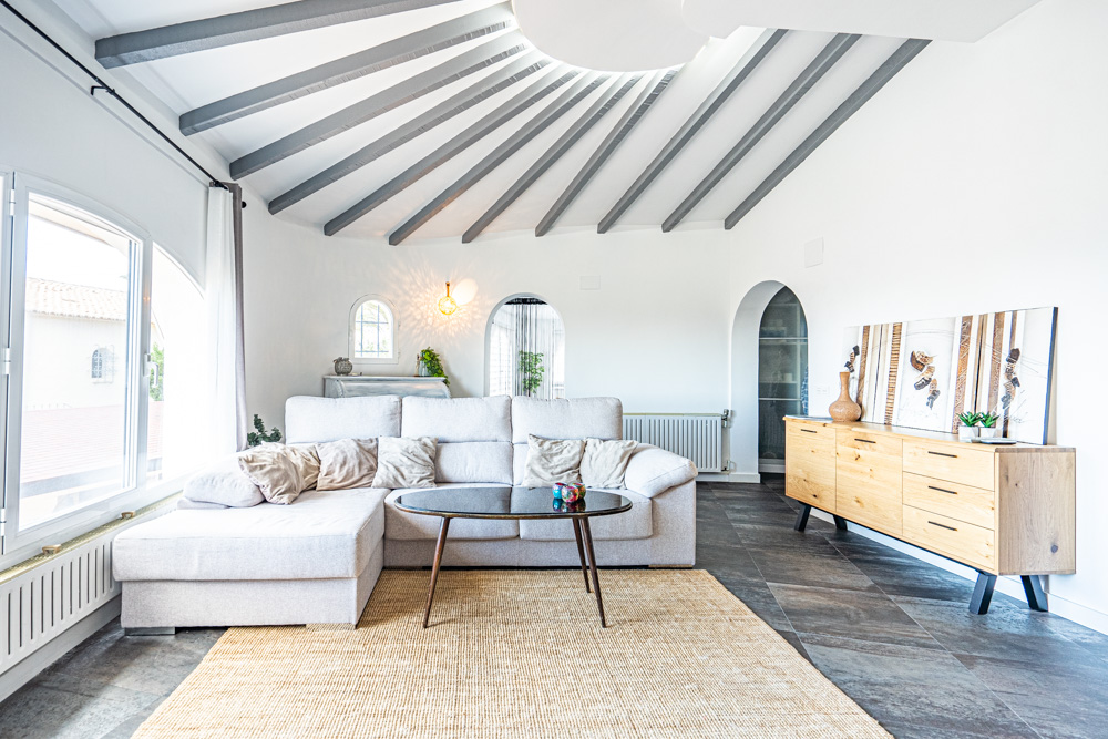 Lichte woonkamer met een radiaal balkenplafond, gebogen witte muren en een donkere tegelvloer. Voorzien van een hoekbank, houten dressoir en grote boogvensters.
