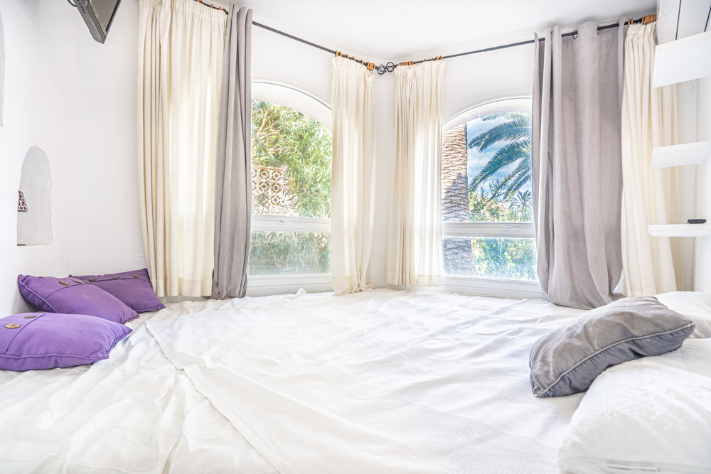 Lichtrijke slaapkamer met gebogen hoekramen die uitkijken op de mediterrane tuin. Voorzien van witte stoffering, paarse accenten en airconditioning.