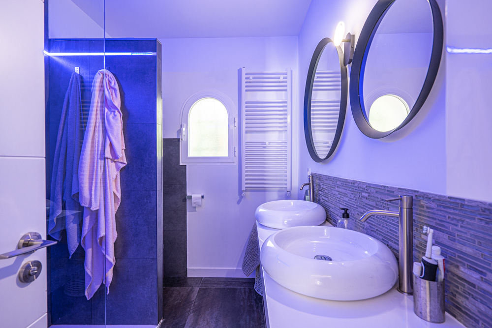 Moderne badkamer met dubbele ronde wastafels, ronde spiegels en een inloopdouche met sfeervolle blauwe LED-verlichting.