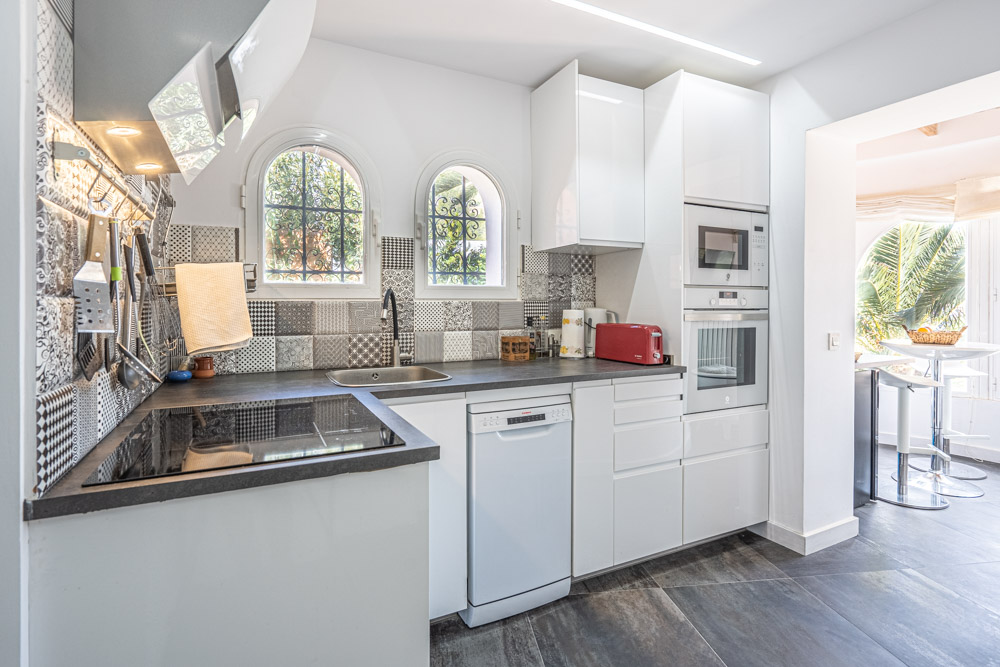 Moderne keuken met witte hoogglanskasten, een decoratieve tegelwand en inbouwapparatuur. Boogvensters zorgen voor natuurlijk licht en uitzicht op de omliggende tuin.