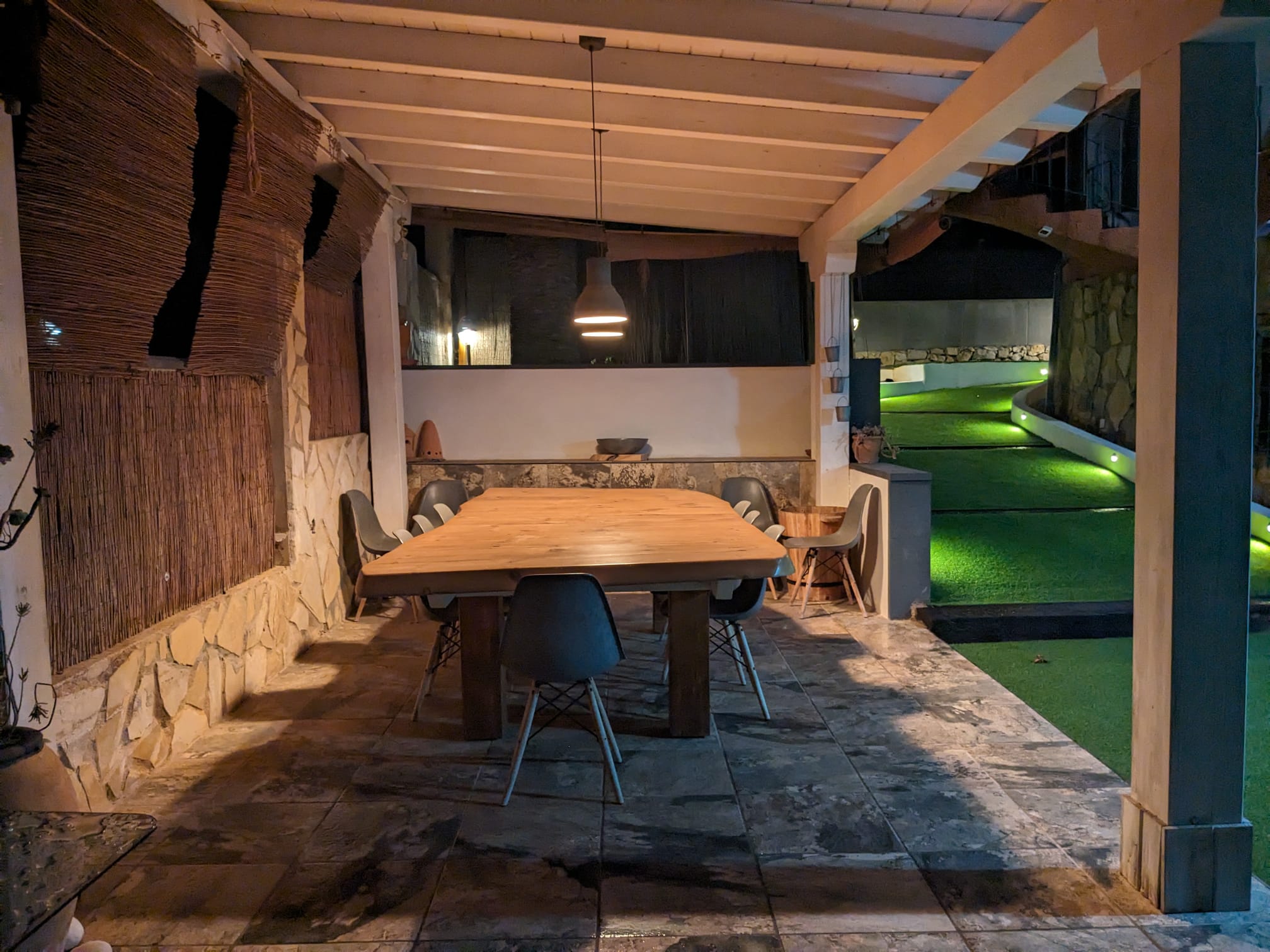 Overdekte buiten-eethoek met een massief houten tafel, natuurstenen wanden en sfeervolle avondverlichting grenzend aan het tuinpad.