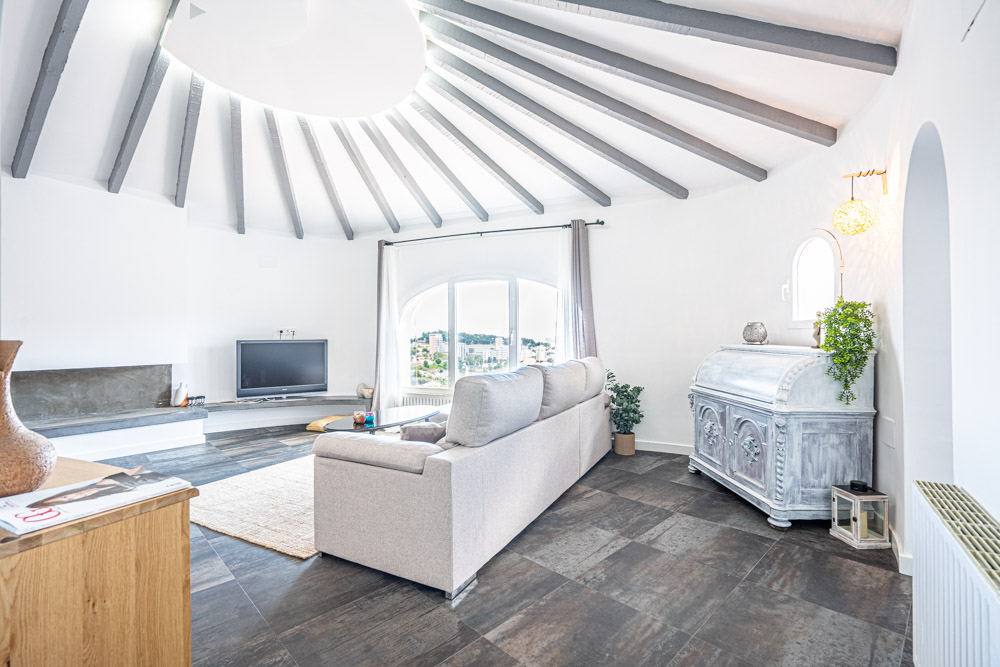 Ronde woonkamer met radiale grijze houten plafondbalken, donkere tegelvloer en een boogvenster met uitzicht op de omgeving van Calpe.