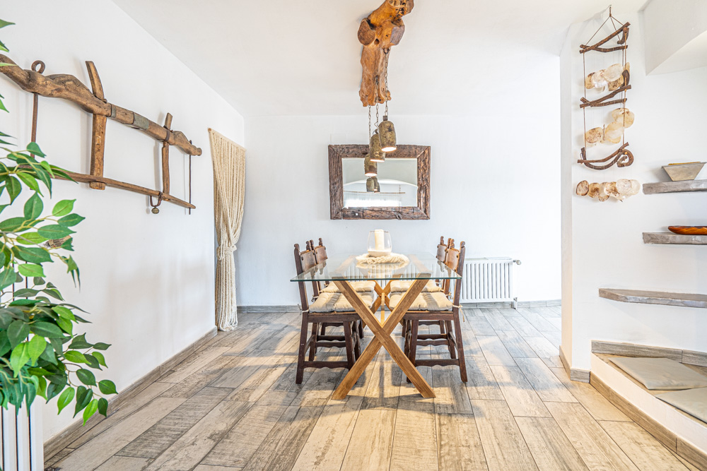 Rustieke eethoek met een glazen tafel en houten onderstel, voorzien van een unieke hanglamp van een natuurlijke boomstam en een grote handgemaakte houten spiegel.
