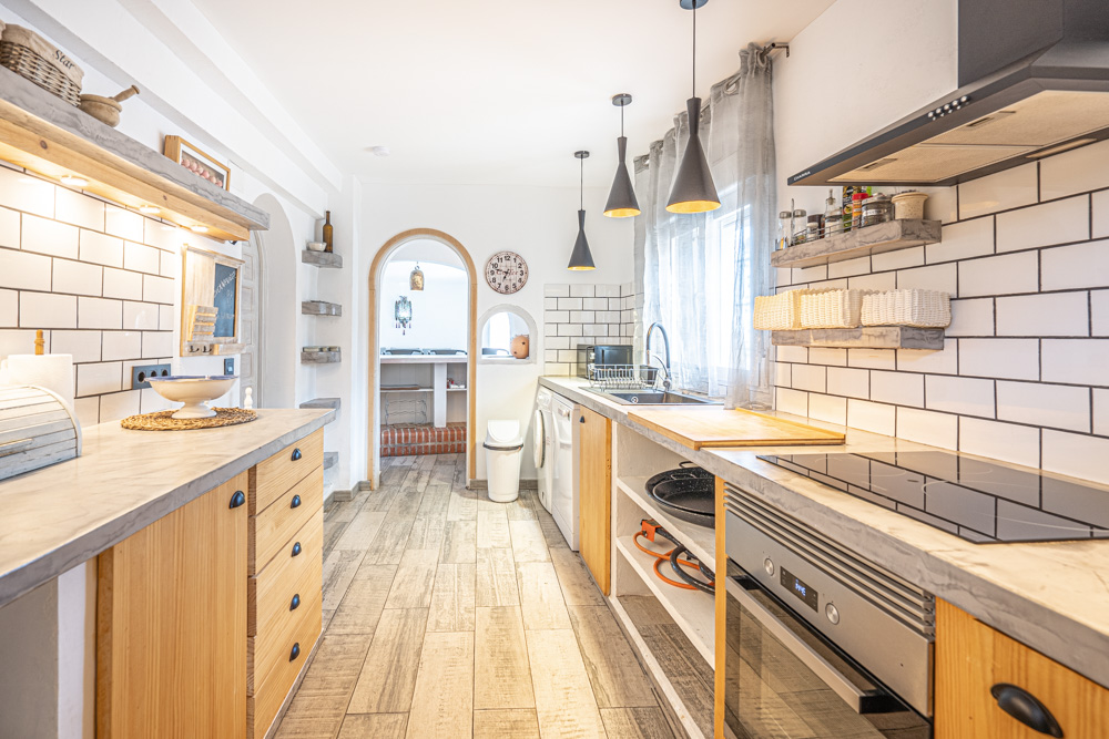 Rustieke keuken met grijze werkbladen, houten kasten en witte metrotegels. Voorzien van inbouwapparatuur, zwarte hanglampen en een boogdoorgang naar de eetkamer.