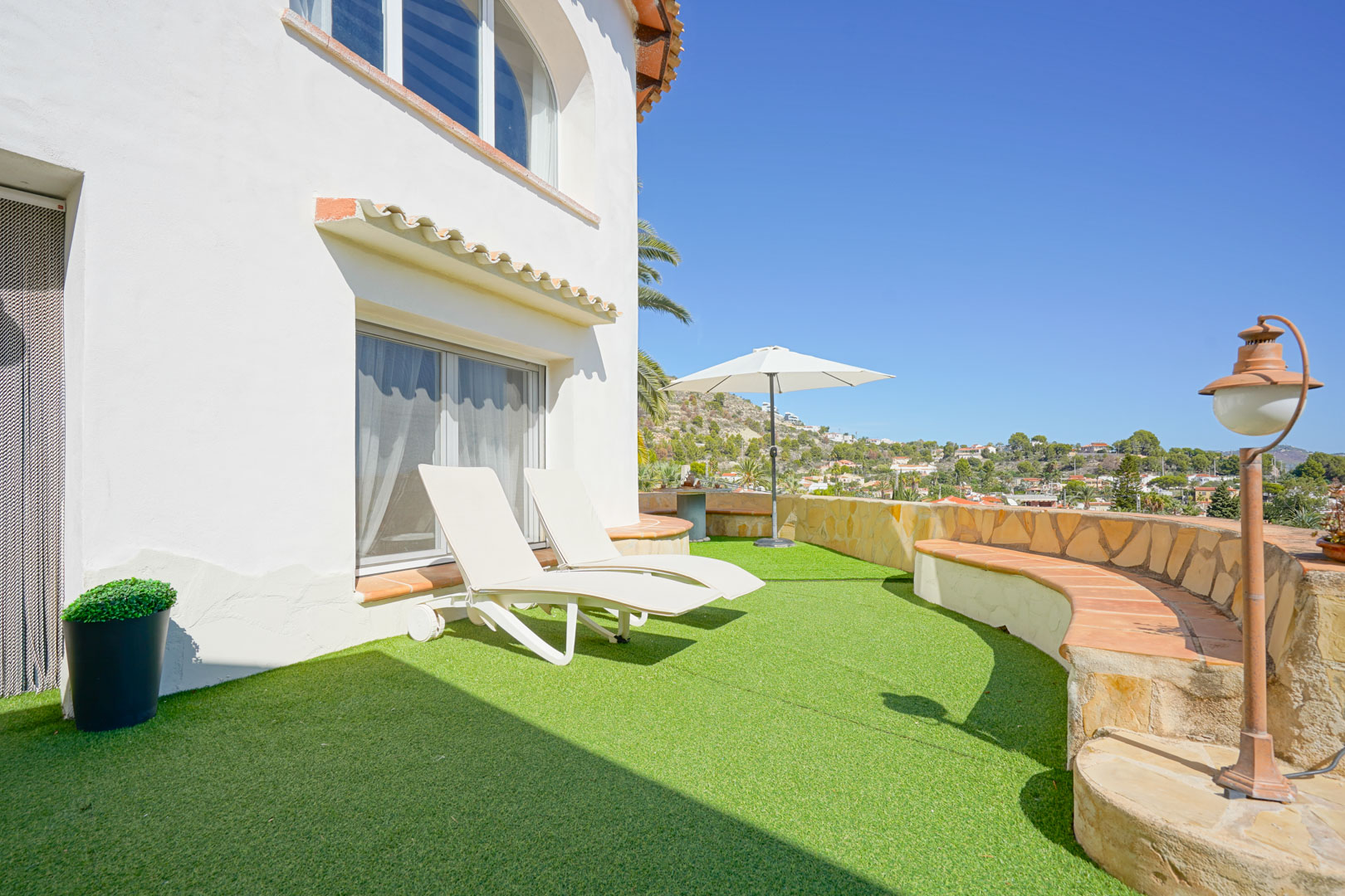 Terras met kunstgras, witte ligbedden en een halfronde stenen muur met ingebouwde zitbank en uitzicht op de omgeving van Calpe.