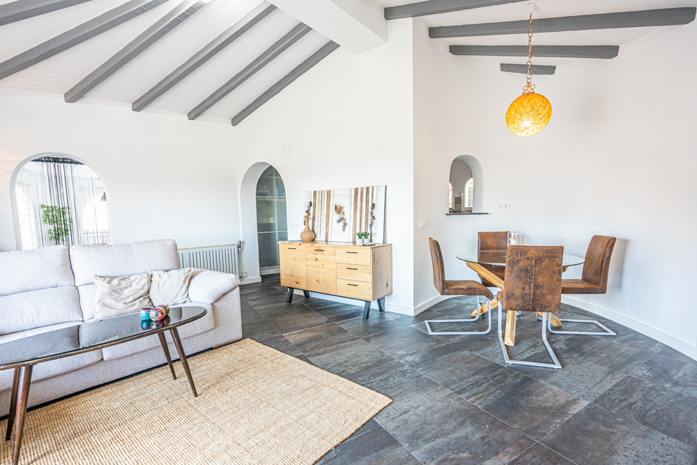 Woon- en eetkamer met grijs geschilderde balkenplafonds, donkere tegelvloer en boogvormige doorgangen. De ruimte is ingericht met een houten dressoir en glazen eettafel.