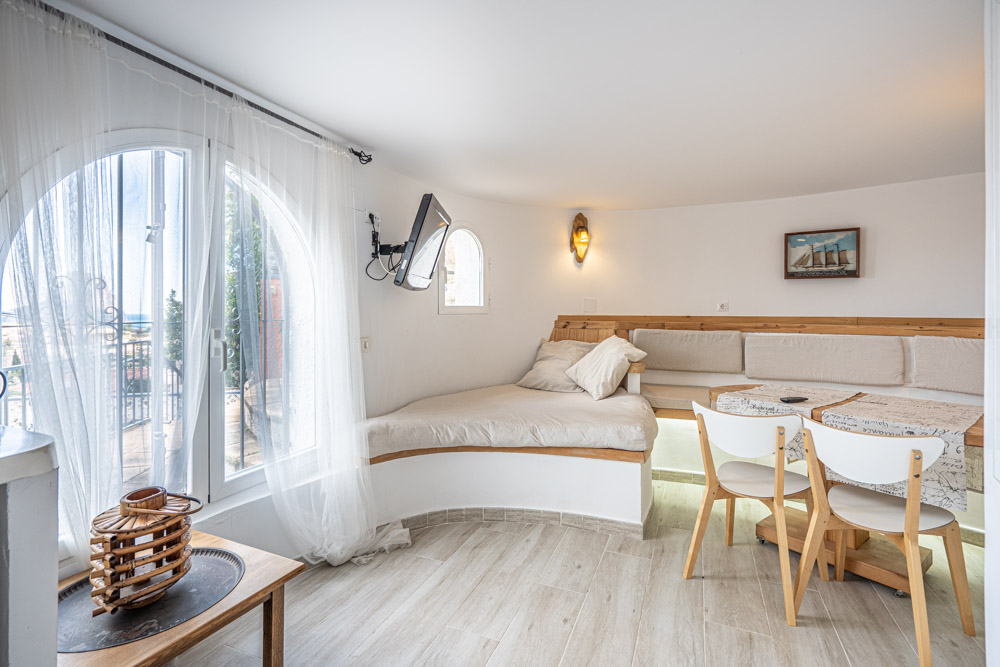 Zithoek met ingebouwde houten bank, eettafel en witte stoelen. De ruimte is voorzien van een boograam en een vloer met houtlook tegels.