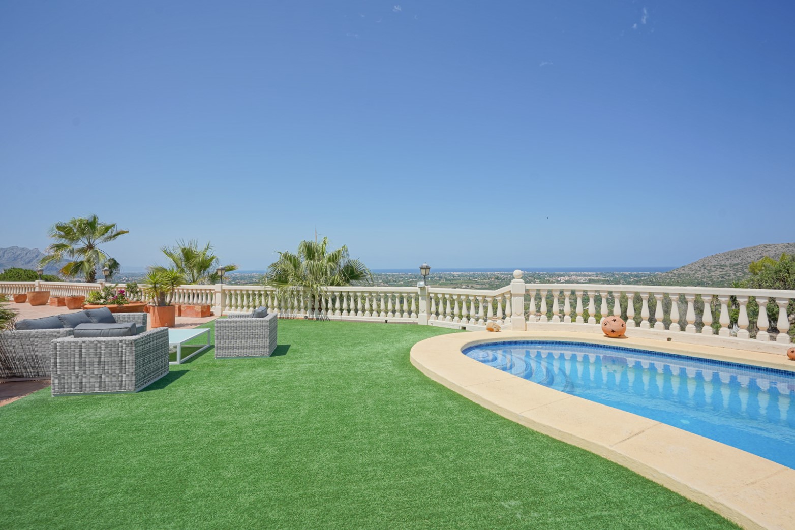 Buitenterras met een niervormig zwembad, kunstgras en wicker loungeset met panoramisch uitzicht over de Middellandse Zee en de vallei van La Sella.