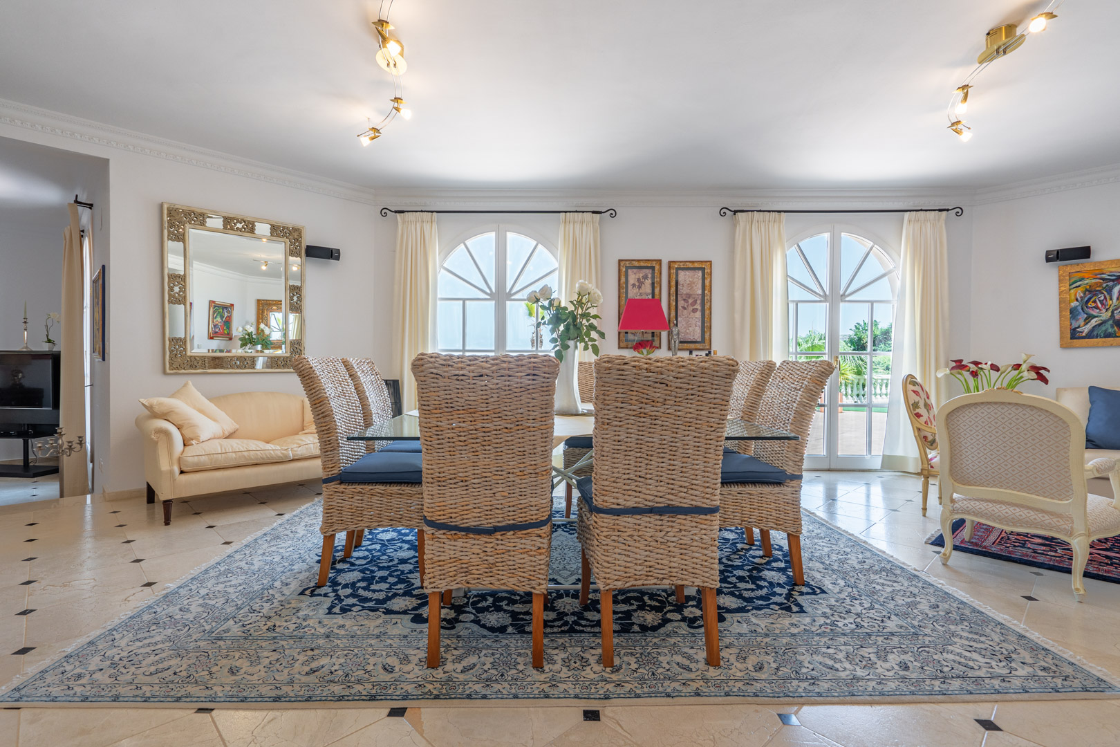 Elegante eetkamer met een glazen tafel en rieten stoelen op een klassiek tapijt. Boogvensters zorgen voor natuurlijk licht en directe toegang tot het terras.