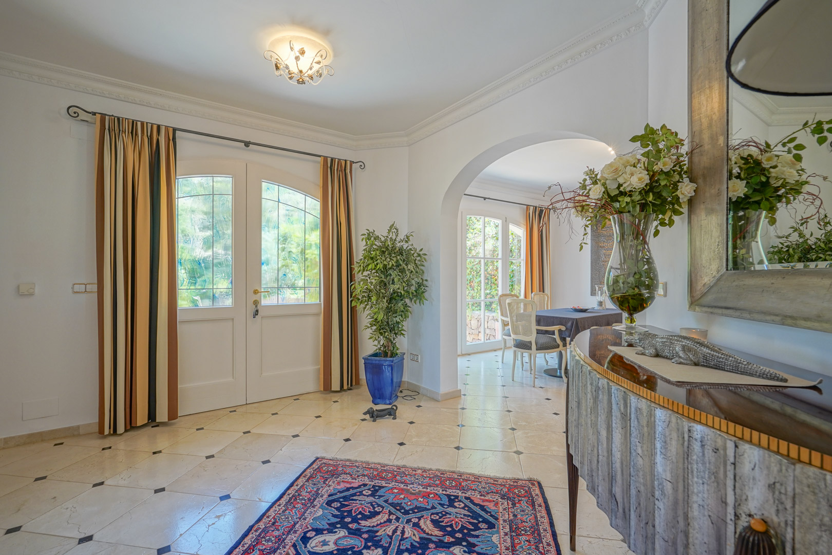 Elegante hal met marmeren vloer en zwarte accenten. Een boogvormige doorgang leidt naar de eetkamer, verlicht door natuurlijk licht via de witte openslaande deuren.