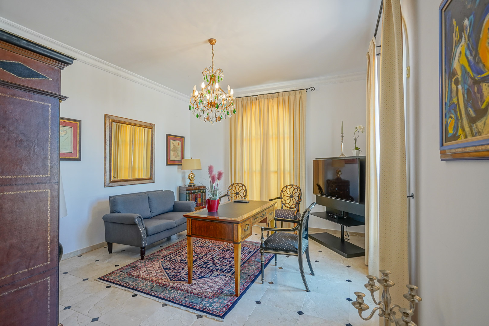 Elegante werkkamer met marmeren vloer, klassiek houten bureau, grijze bank en een kristallen kroonluchter met groene details.