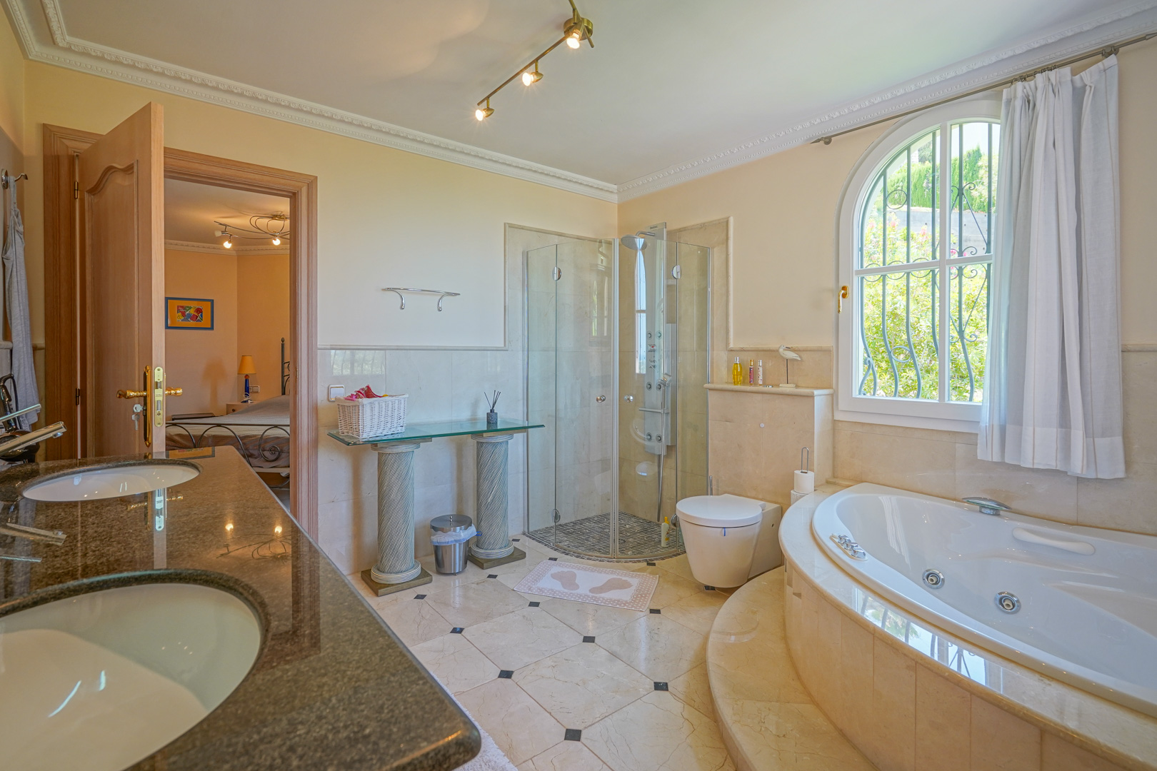 En-suite badkamer met rond hydromassagebad, glazen douchecabine en dubbele granieten wastafel. Voorzien van marmeren vloeren en een boogvenster met uitzicht op de tuin.