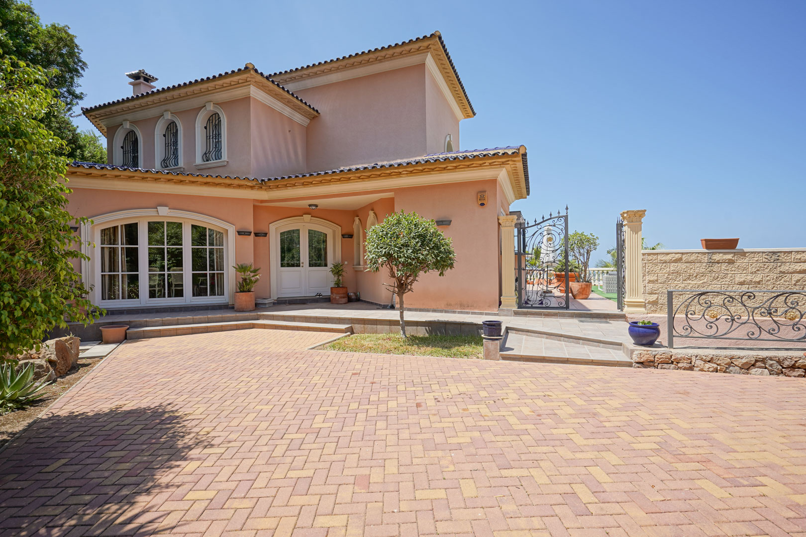 Exterieur van de villa met een oprit in visgraatmotief, boogramen met smeedijzeren details en een witte dubbele voordeur tegen een helderblauwe lucht.