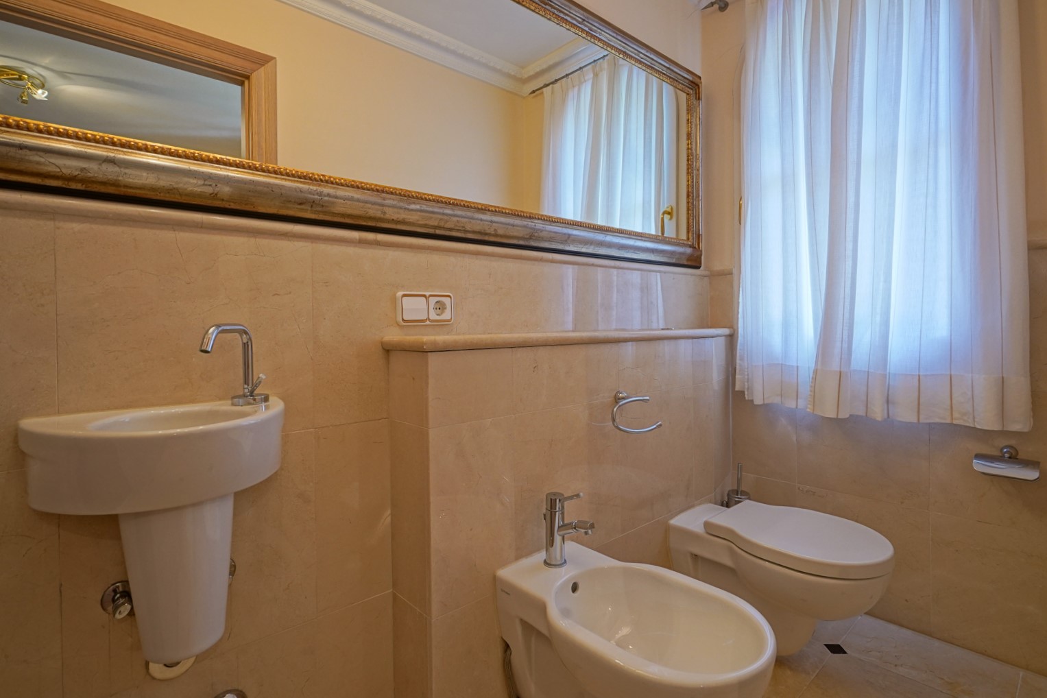 Gastentoilet met crème marmeren wanden, voorzien van een wastafel, bidet en toilet. Een grote spiegel met sierlijst en natuurlijk licht via het raam creëren een ruimtelijk effect.