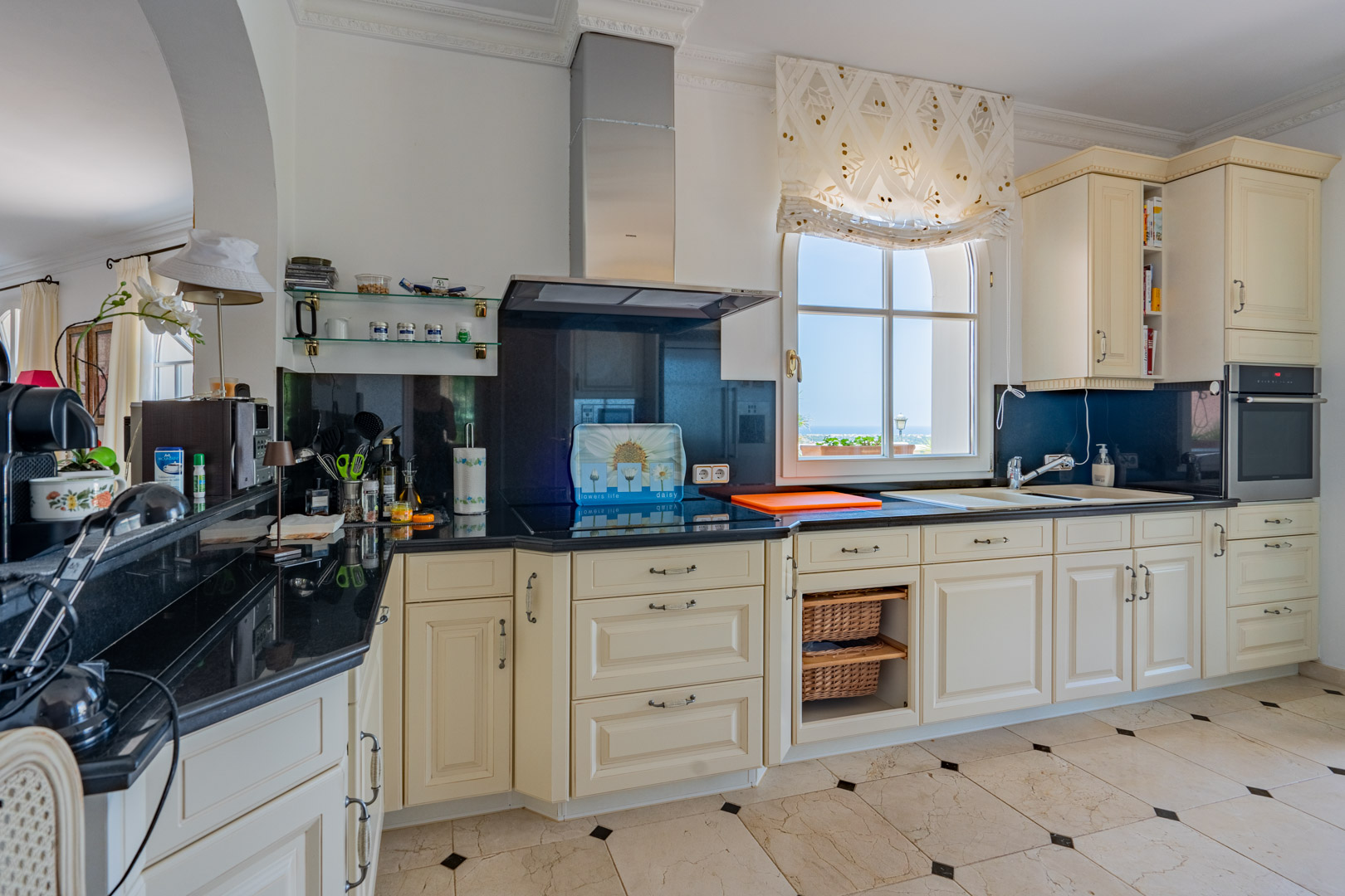 Klassieke keuken met crèmekleurige kasten, zwart granieten werkbladen en marmeren vloer. Voorzien van inbouwapparatuur en een venster met uitzicht op de kustlijn.