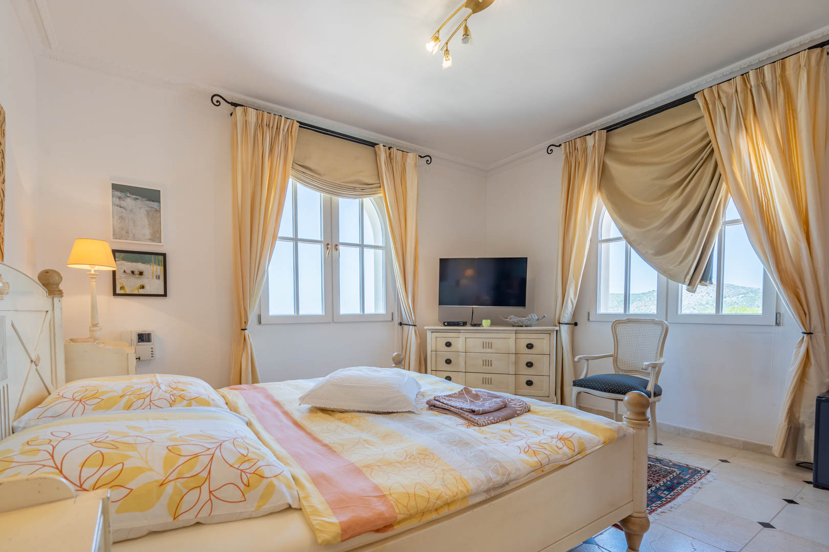 Lichte slaapkamer met wit houten meubilair, boogvensters met crème gordijnen en een tegelvloer met donkere accenten. Voorzien van een tv-meubel en een klassieke fauteuil.