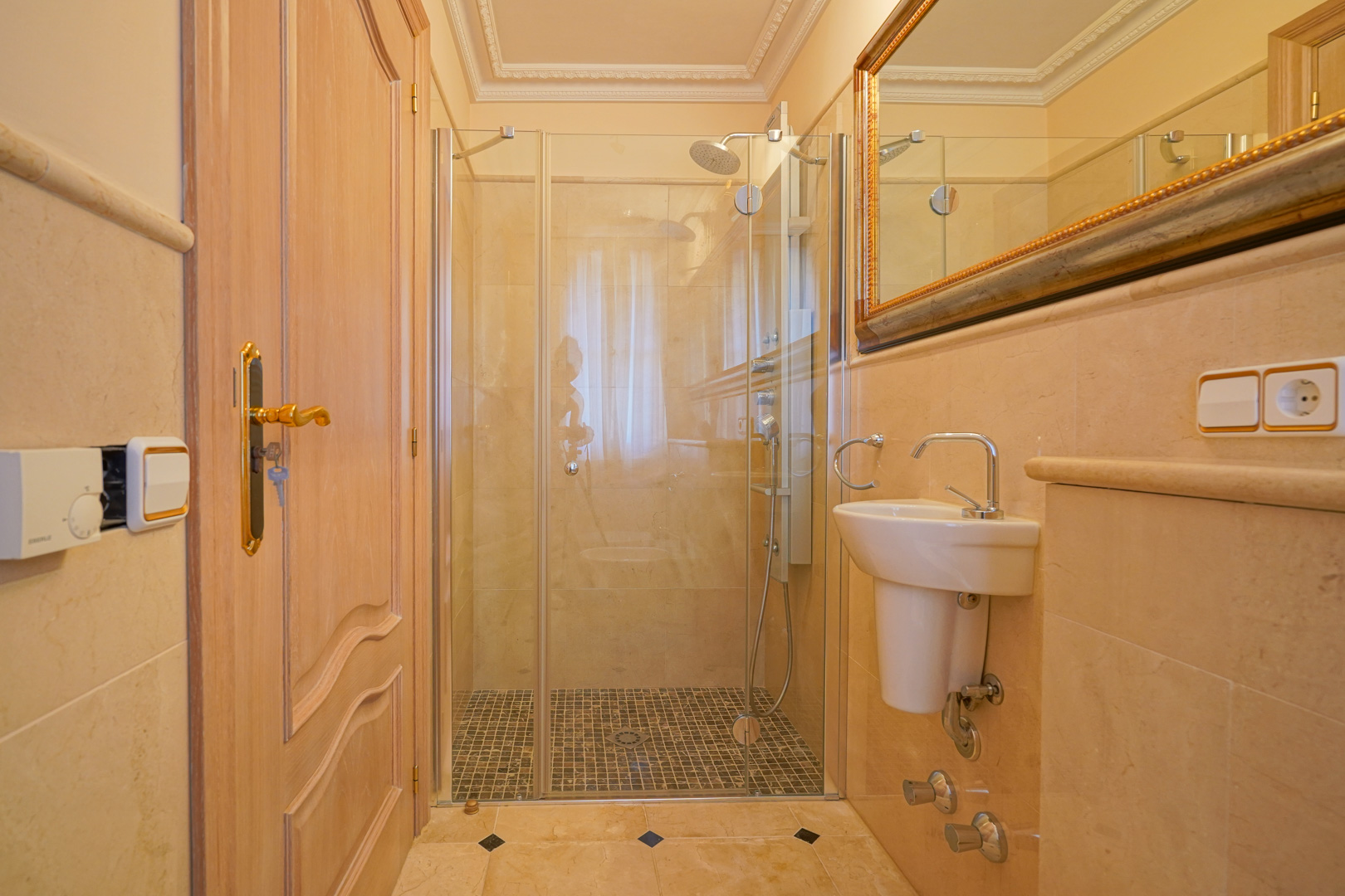 Met marmer afgewerkte badkamer voorzien van een inloopdouche met mozaïekvloer en glazen wand. Inclusief een compacte wastafel en een spiegel met gouden lijst.