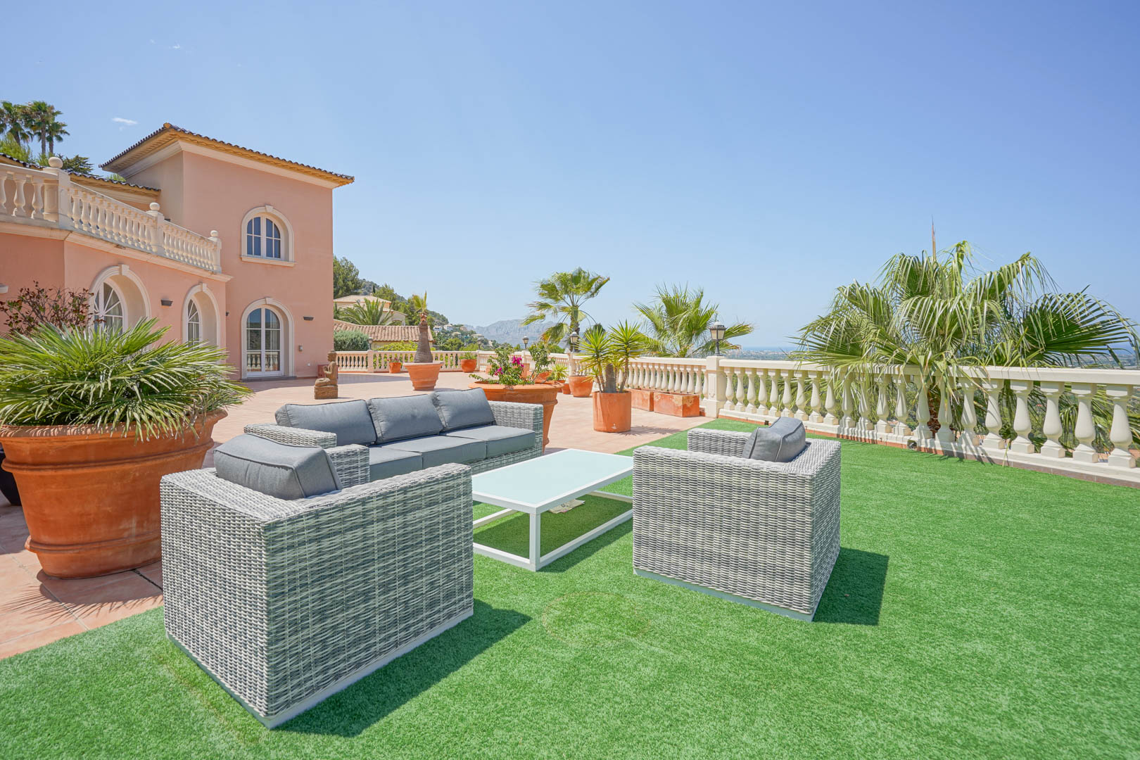 Ruim terras met grijze wicker loungeset op kunstgras, omgeven door terracotta potten en een panoramisch uitzicht over de vallei en de kustlijn.