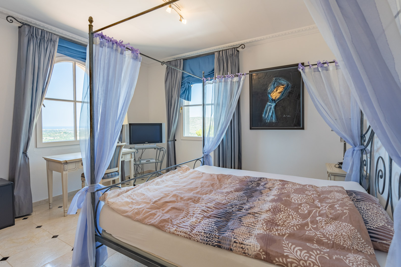 Slaapkamer met metalen hemelbed en lichte gordijnen, marmeren vloer met zwarte details en een werkplek bij het raam met uitzicht op de vallei.