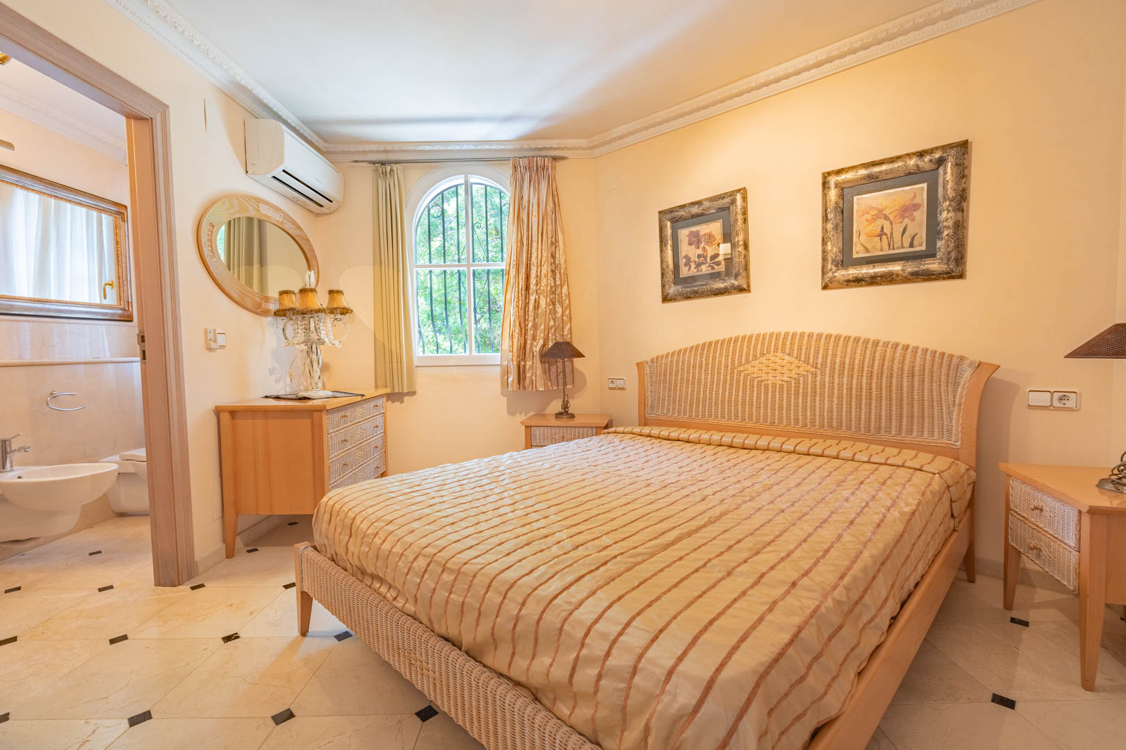 Slaapkamer met rotan bed, marmeren vloer met zwarte accenten en aangrenzende badkamer. Voorzien van airconditioning, sierlijsten en een klassiek boogvenster.