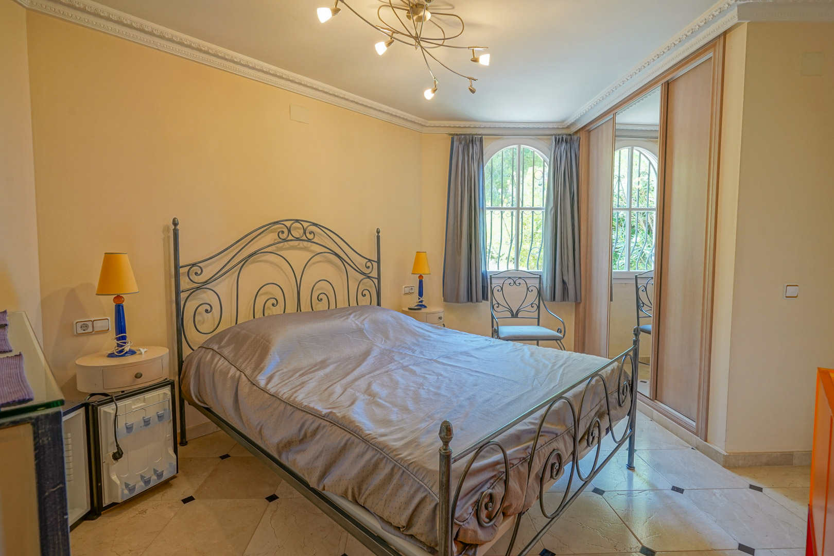 Slaapkamer met smeedijzeren bed, marmeren vloer met zwarte details, inbouwkasten en een karakteristiek boograam met zicht op de tuin.
