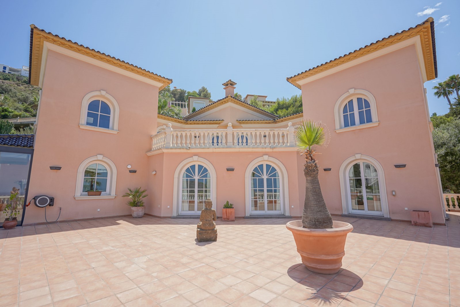 Symmetrische zalmkleurige gevel met boogvensters, een boventerras met witte balustrades en een ruime binnenplaats met terracotta tegels en decoratieve elementen.