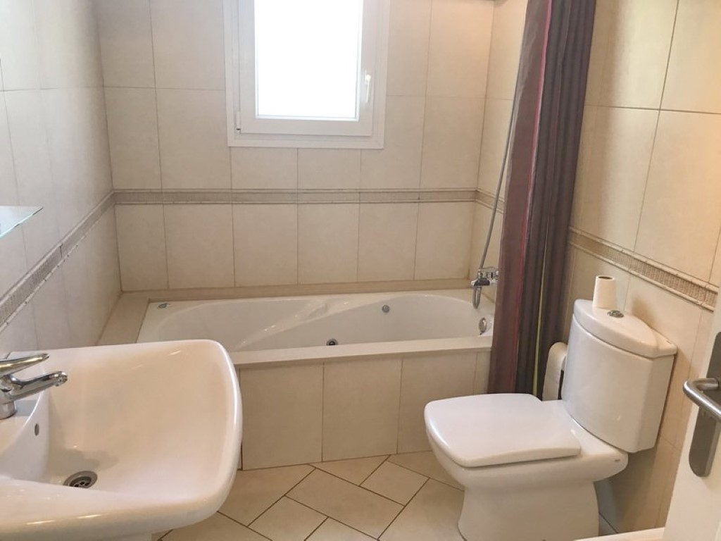 Badkamer voorzien van een ingebouwd hydromassagebad, wit voetstuk wastafel en toilet. Afgewerkt met neutrale wandtegels, een decoratieve rand en natuurlijk lichtinval.