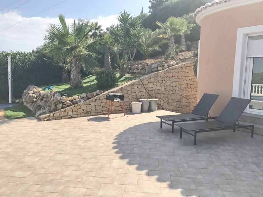 Betegeld terras met ligstoelen en barbecue, grenzend aan een natuurstenen stapelmuur en een aangelegde tuin met palmbomen op een helling.