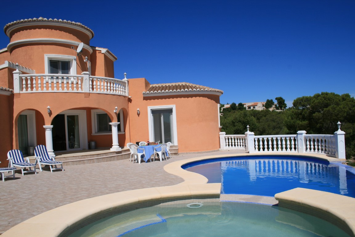 Buitenaanzicht van een mediterrane villa met niervormig zwembad, geïntegreerde jacuzzi en een ruim betegeld terras met witte balustrades en boogarchitectuur.