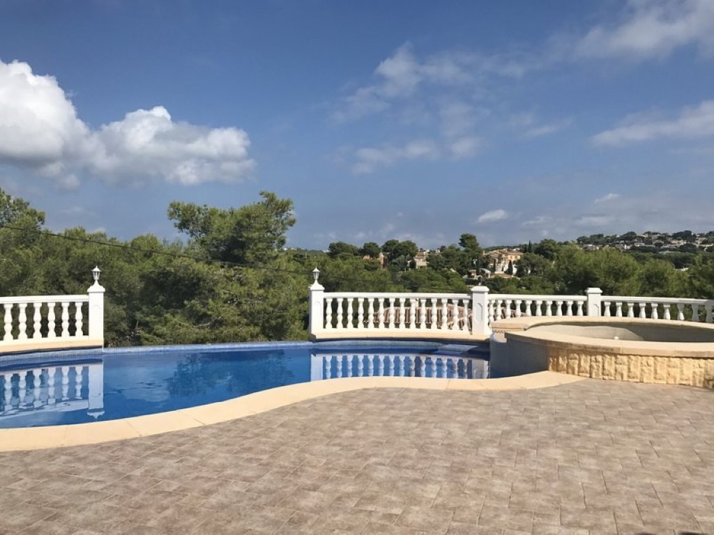 Gebogen zwembad met aangrenzende jacuzzi, omgeven door een betegeld terras en witte balustrade met uitzicht op de pijnbomen en heuvels van Javea.