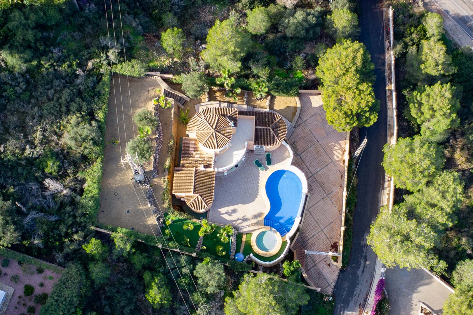 Luchtfoto van de villa met terracotta pannendaken, een niervormig zwembad en een ronde jacuzzi, omgeven door een dicht mediterraan boslandschap.
